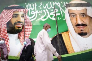 People wearing face masks walking past a banner of Saudi King Salman and Crown Prince Mohammed bin Salman