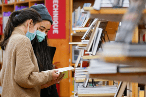 On left: two masked individuals looking down at books in a bookstore; books and bookshelves in background