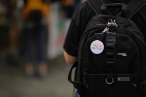 a student wears a backpack with a pin that reads "Facts Matter"