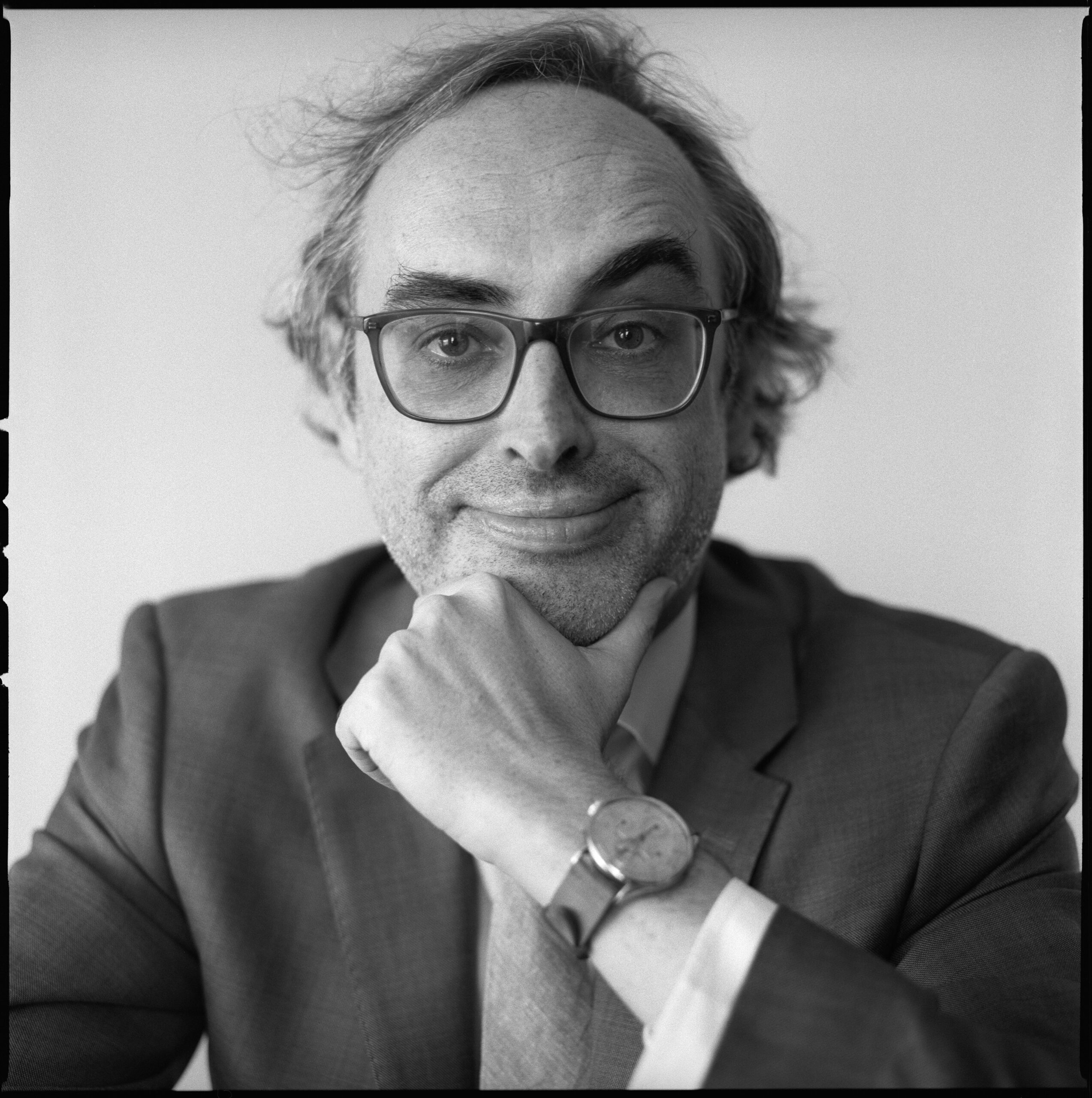 A man in a suit and glasses sits with his chin resting on his hand, looking at the camera and smiling slightly. He is wearing a wristwatch and has tousled hair. The photo is black and white.