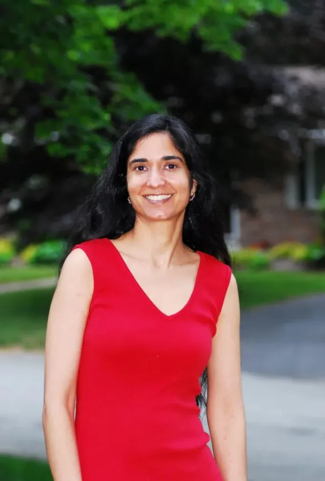 A woman with long black hair, wearing a sleeveless red dress, stands outdoors smiling. Green trees and blurred buildings are in the background.