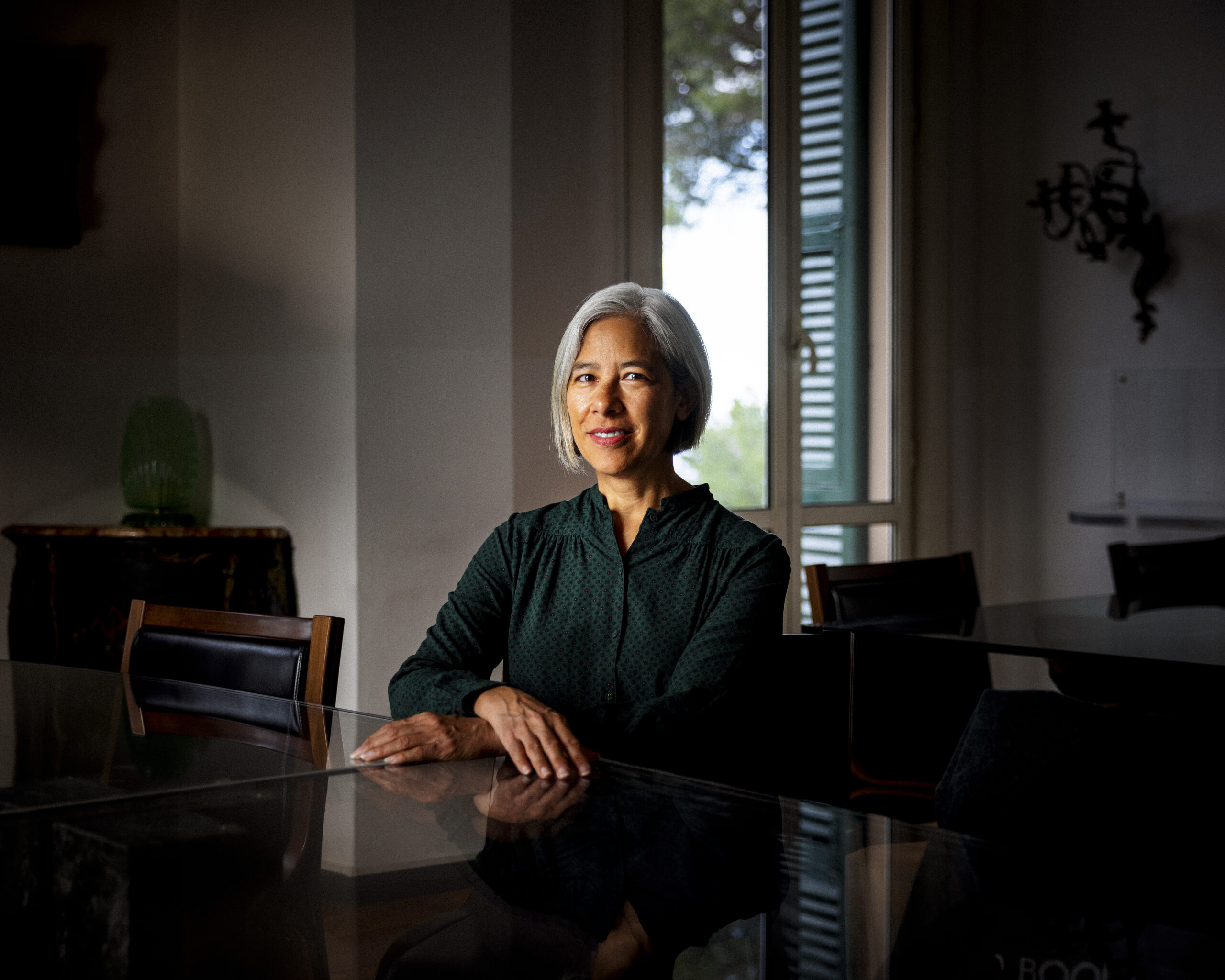 A woman with short gray hair, wearing a dark green blouse, sits at a reflective table in a dimly lit room with large windows and soft natural light. She is smiling and looking slightly to the side.