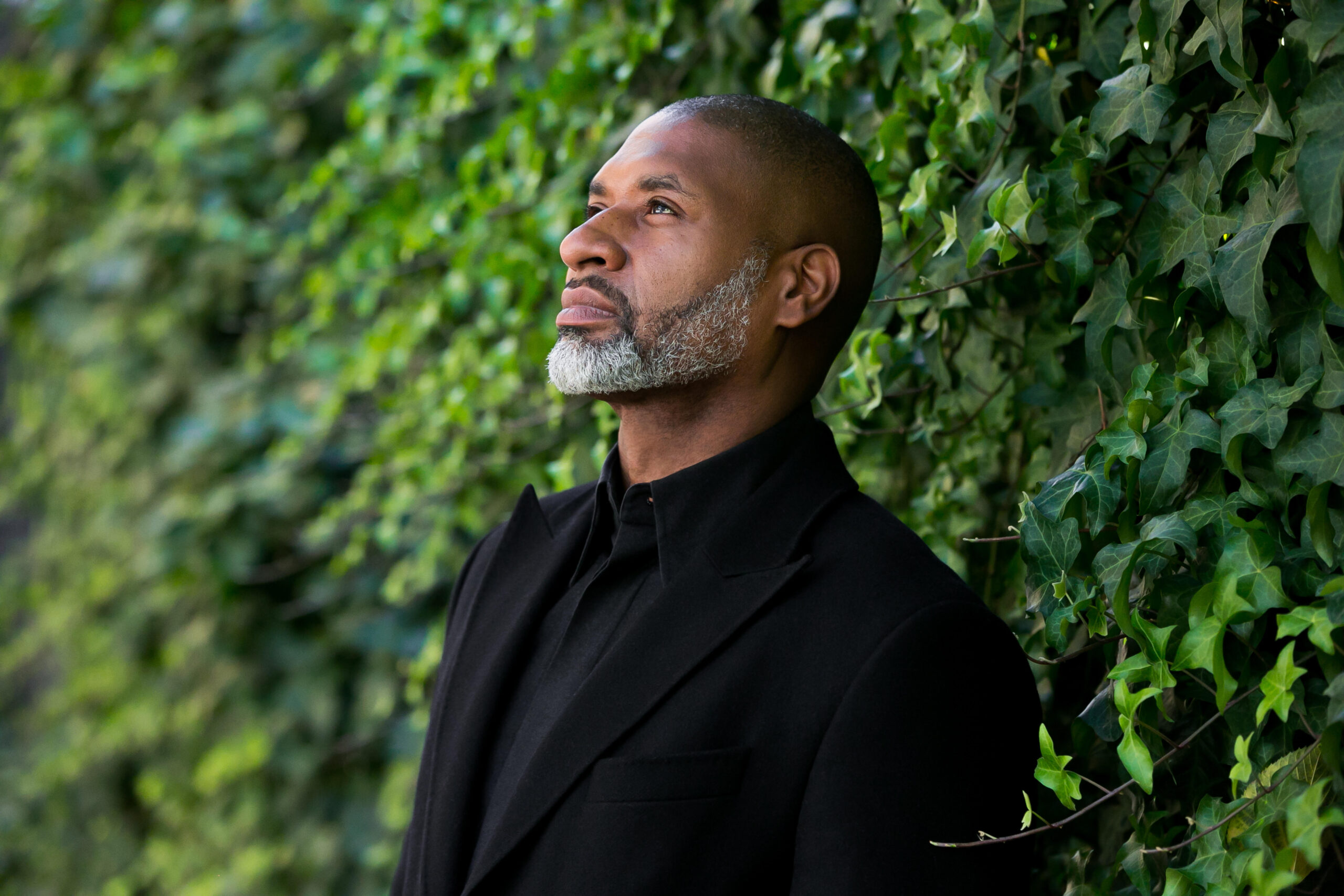 A man with a salt-and-pepper beard and mustache, wearing a black coat, stands against a leafy green wall, looking thoughtfully upward.