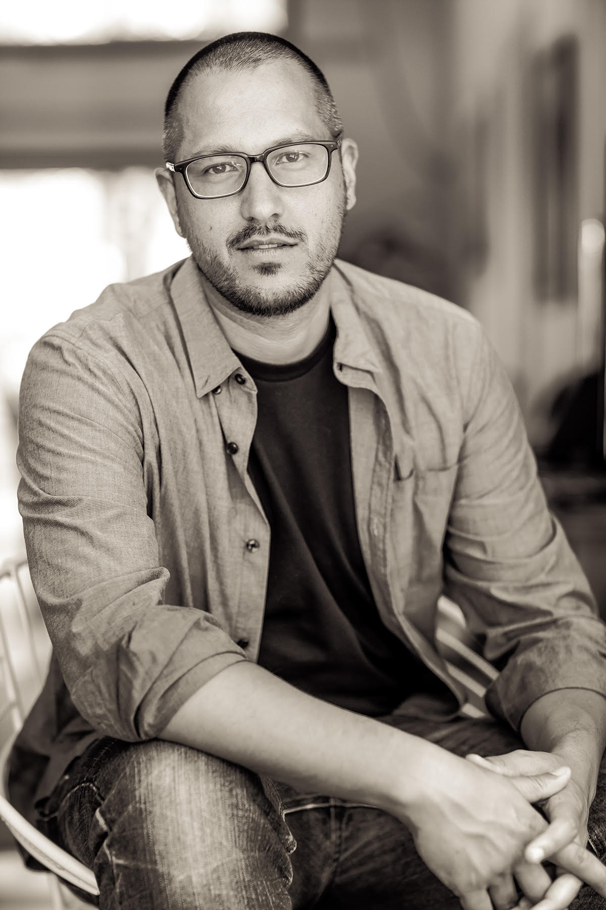 A man with glasses and a beard sits in a chair indoors, wearing a light button-up shirt over a dark t-shirt and jeans. He looks directly at the camera with a neutral expression. The background is softly blurred. Black and white image.