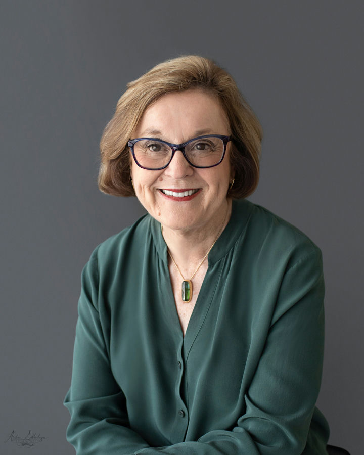 A woman with brown hair and glasses smiles while wearing a green blouse and necklace against a gray background.