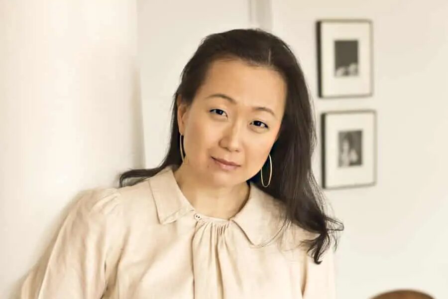 A person with long dark hair, wearing a beige blouse and hoop earrings, leans slightly against a white wall. Two framed pictures are visible in the blurred background.