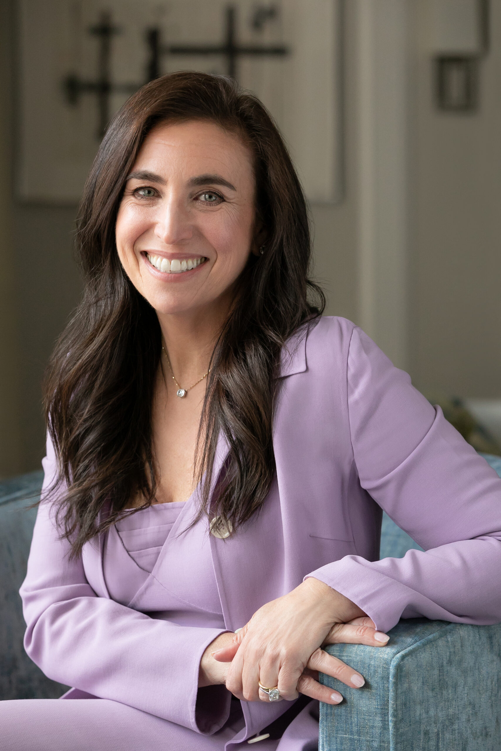 Smiling woman with long dark hair wearing a lavender suit, seated with hands resting on a blue chair. Background features abstract black and white artwork.