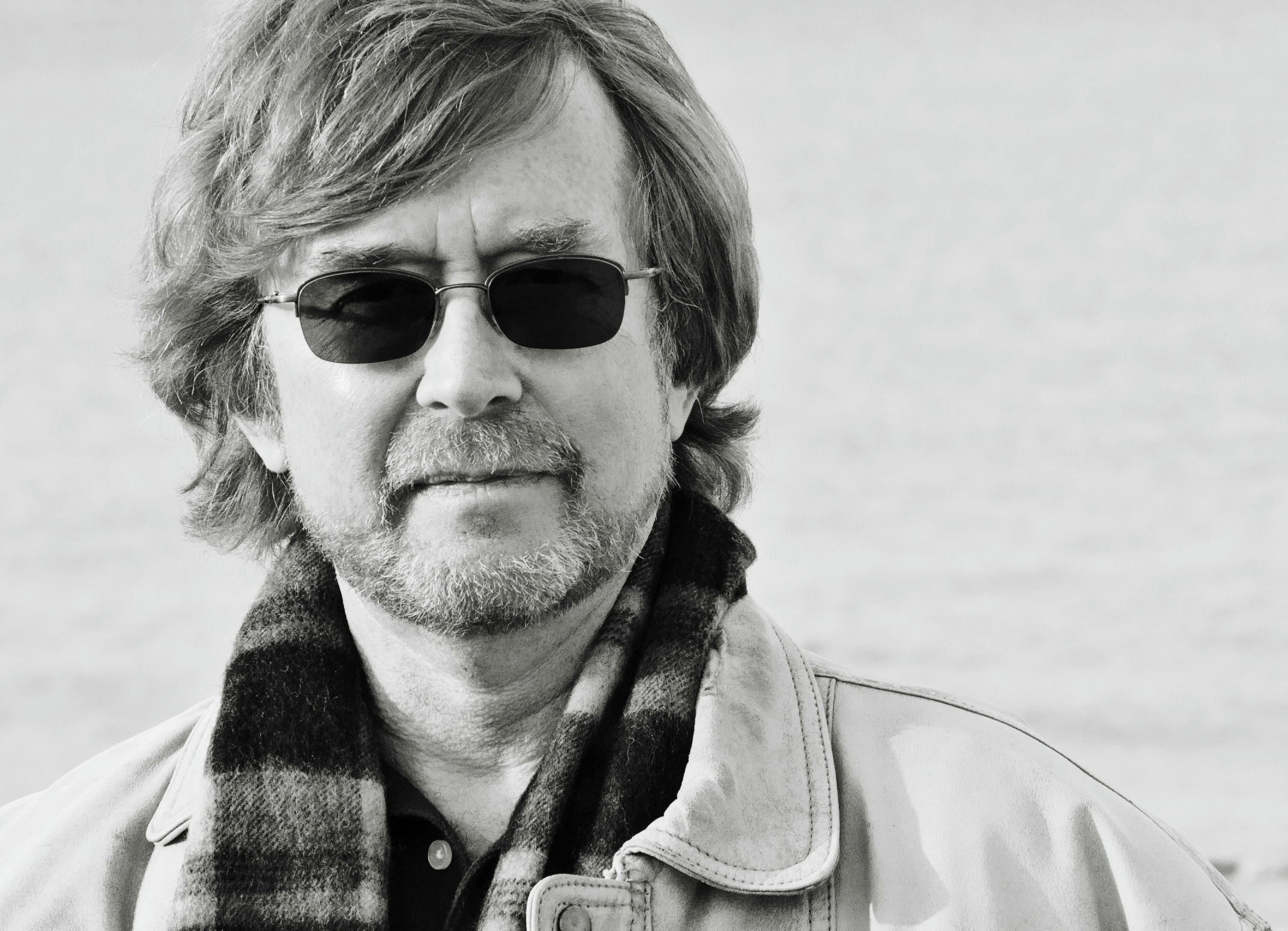 A man with wavy hair and a beard wearing sunglasses, a plaid scarf, and a light-colored jacket. He stands against a blurred background, which appears to be a large body of water. The photo is in black and white.