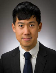 A man with short black hair wearing a black suit, white shirt, and dark tie poses for a professional headshot against a gray background.