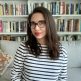 Alia Hanna Habib headshot: Alia sits in front of a white shelf full of books. She is wearing a shirt with horizontal black stripes and dark framed glasses.