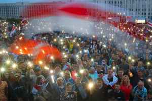 Belarusian protesters holding up their light-up phones, with faded, old Belarusian national flag on top of image