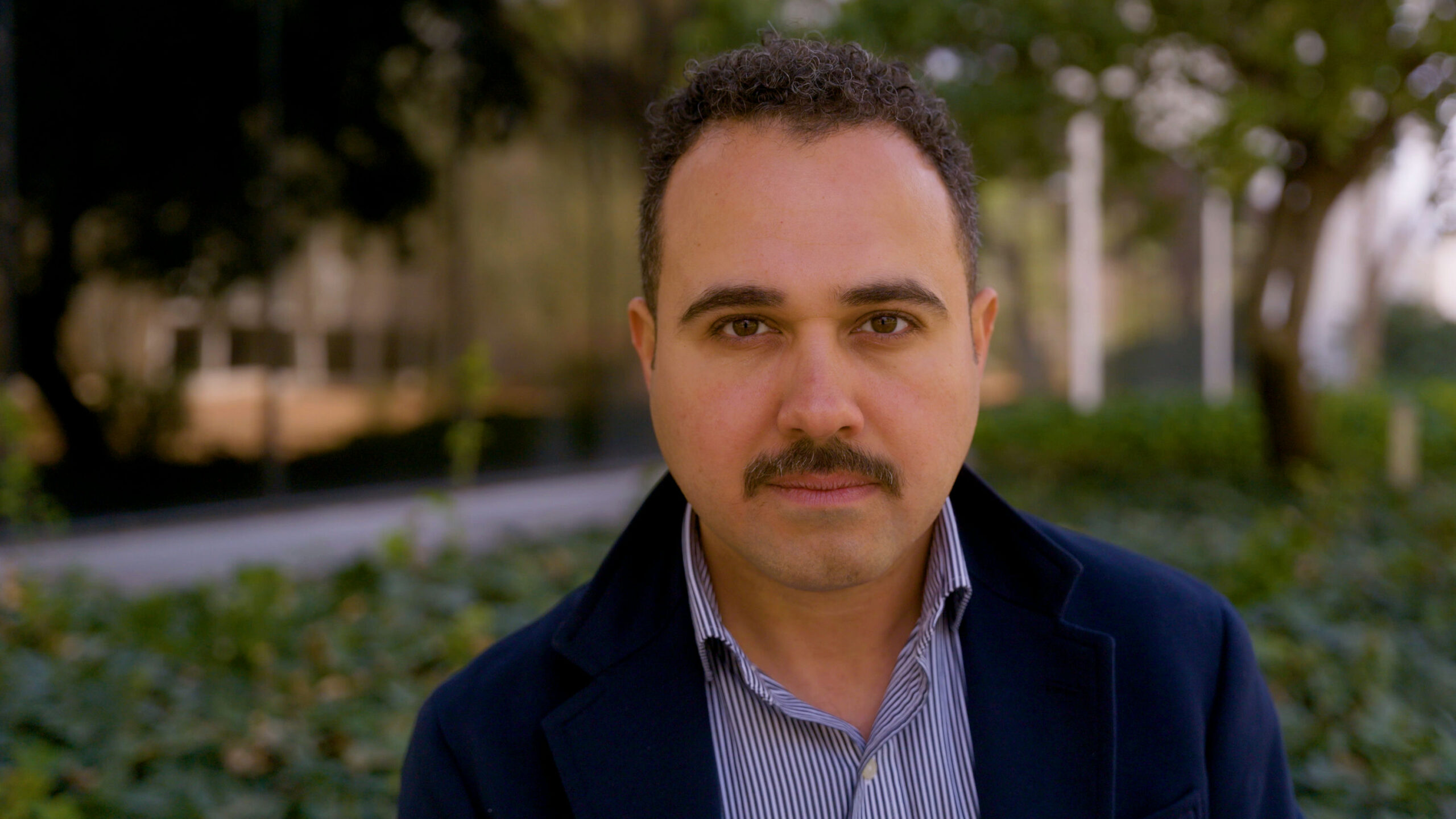 A person with a mustache and short curly hair is wearing a dark jacket over a striped shirt. They are standing outdoors with greenery and blurred trees in the background.