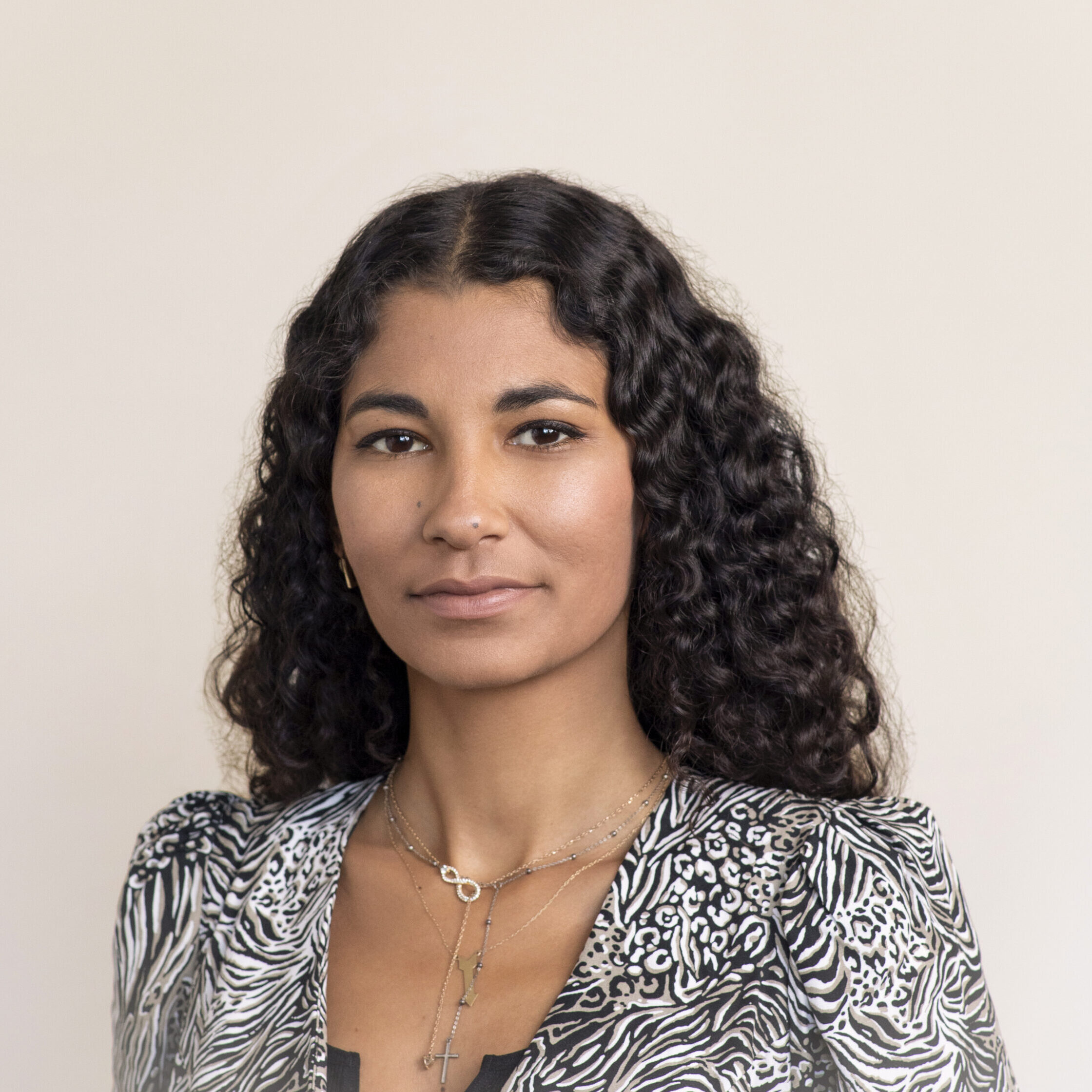 A person with long, curly hair poses for a portrait against a neutral background. They are wearing a patterned blouse, layered necklaces, and have a calm expression.