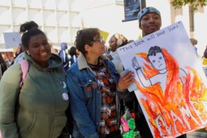 Three students stand at a rally in New Haven, CT. One student holds a hand drawn sign featuring the cover of Flamer by Mike Curato. The students are smiling.