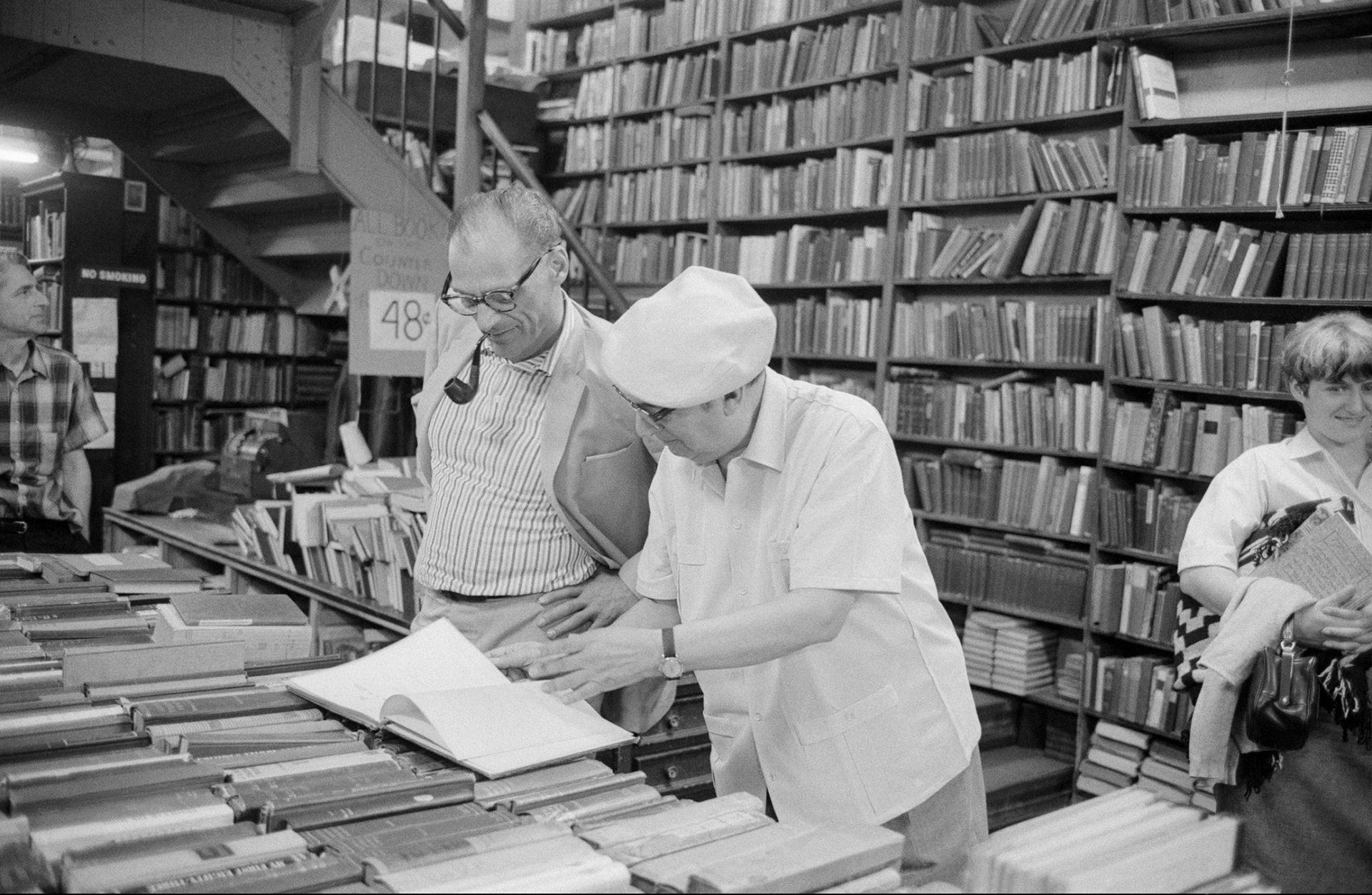 Two people stand by a table covered with books in a library or bookstore, looking at an open book together—perhaps exploring PEN America history. Shelves filled with books line the walls, and a few others are browsing nearby.