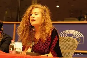 A young woman with curly blonde hair speaks into a microphone while seated at a table. She wears a patterned red and black top, with a European Parliament emblem visible in the background.