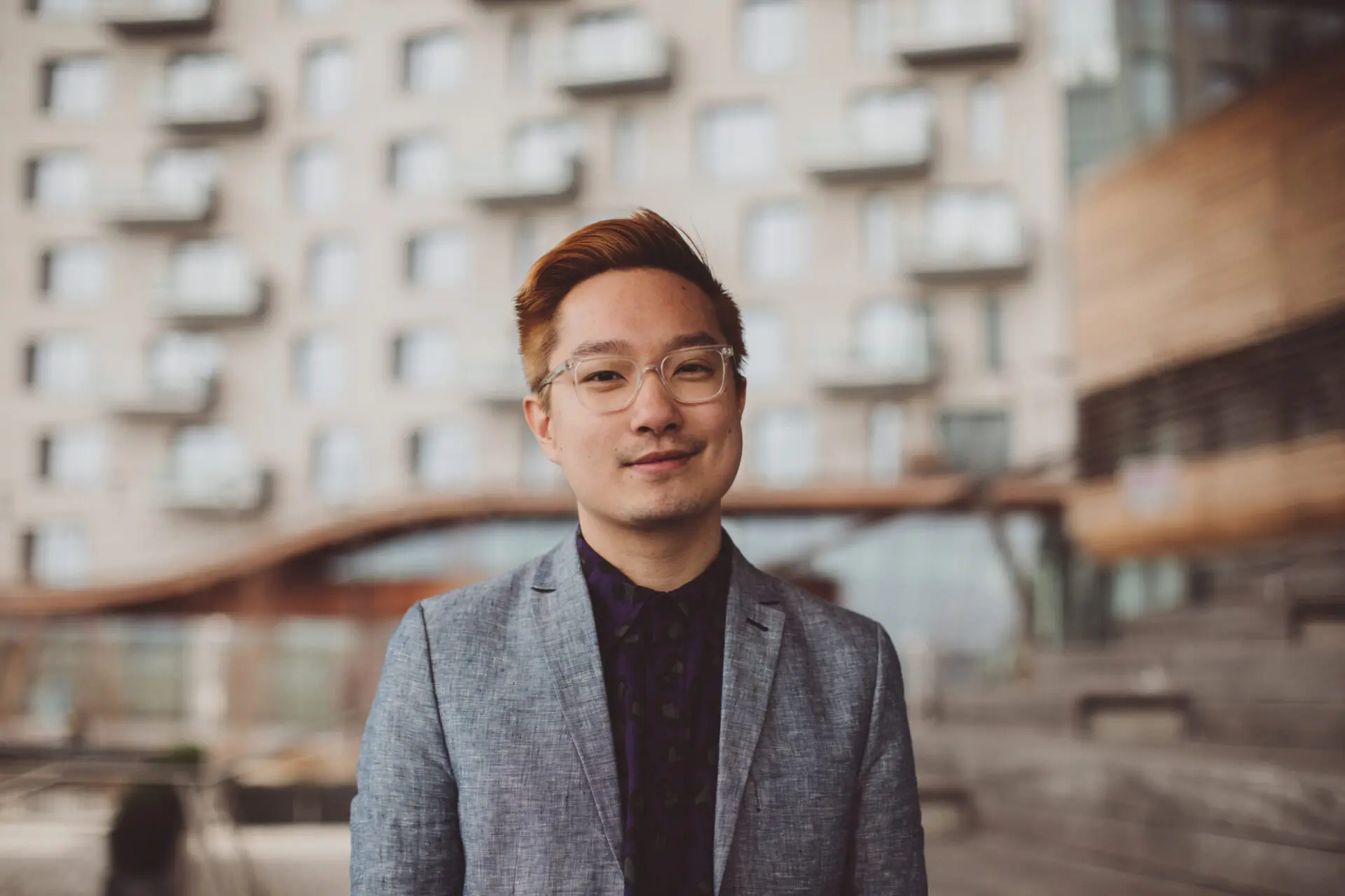 A person wearing glasses and a gray blazer stands outdoors in front of a modern building with balconies, looking at the camera and smiling softly. The background is slightly blurred.