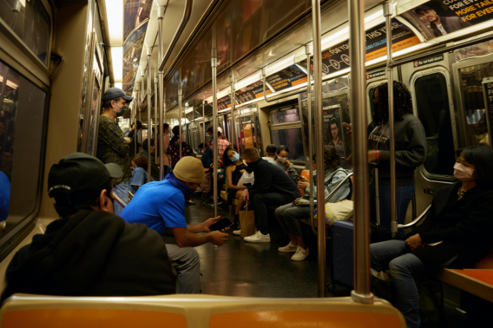 Passengers sit and stand inside a subway car. Most people are wearing masks, some are looking at phones, and the seats are orange and beige. Overhead, advertisements line the walls of the train.