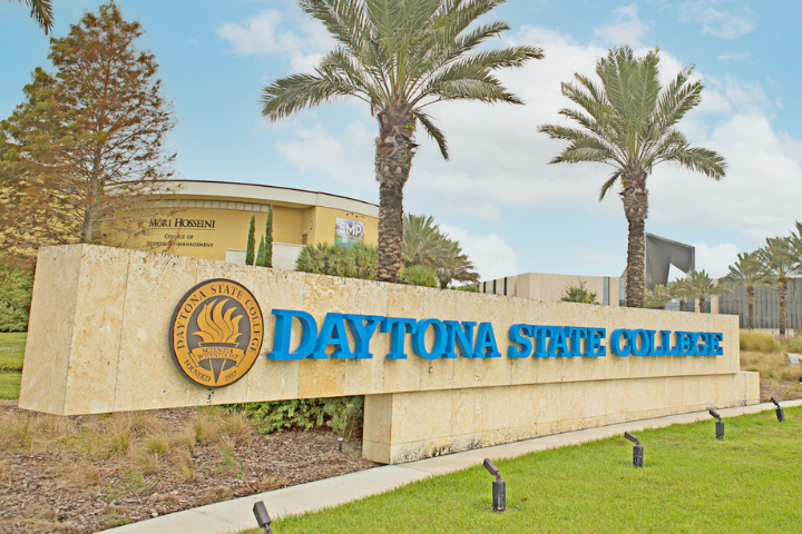 Large stone sign for Daytona State College with the college seal, surrounded by palm trees and grass, with campus buildings visible in the background under a partly cloudy sky.
