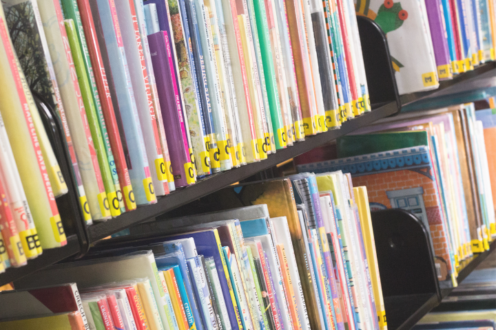 Colorful books are neatly arranged on two tilted shelves in a library, with yellow and black labels on their spines. The books are mostly upright, and their covers and spines display a variety of patterns and colors.