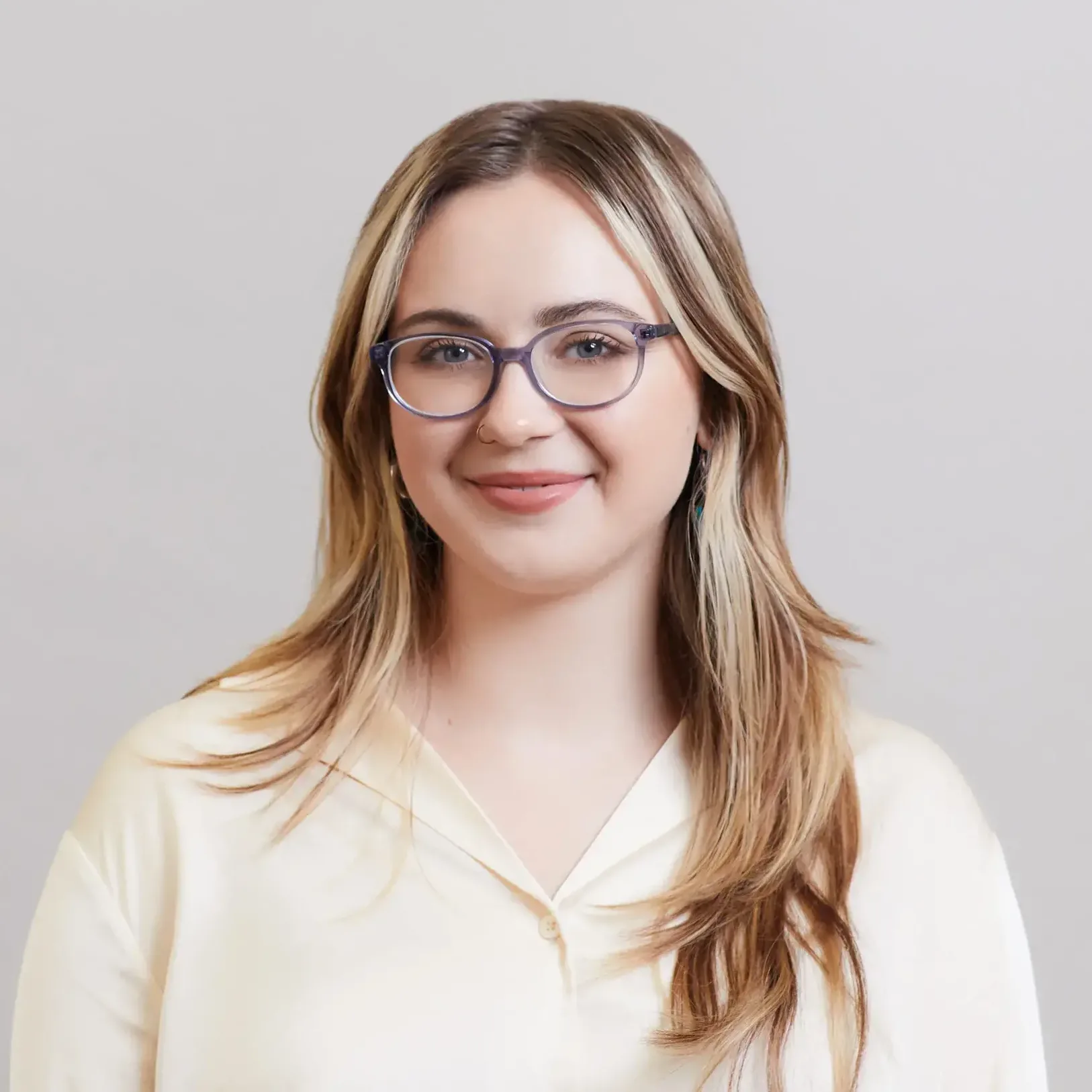 A young woman with long, light brown hair and glasses smiles softly. She is wearing a cream-colored blouse and stands in front of a plain, light gray background.