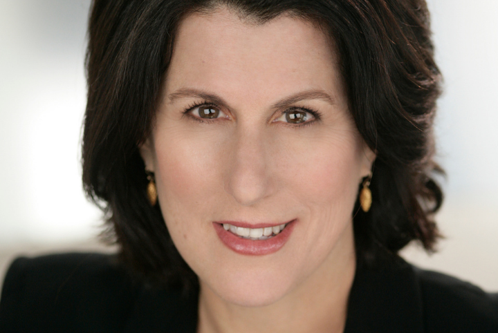 A woman with shoulder-length dark brown hair, wearing gold earrings and a black top, smiles slightly while looking directly at the camera. The background is softly blurred.