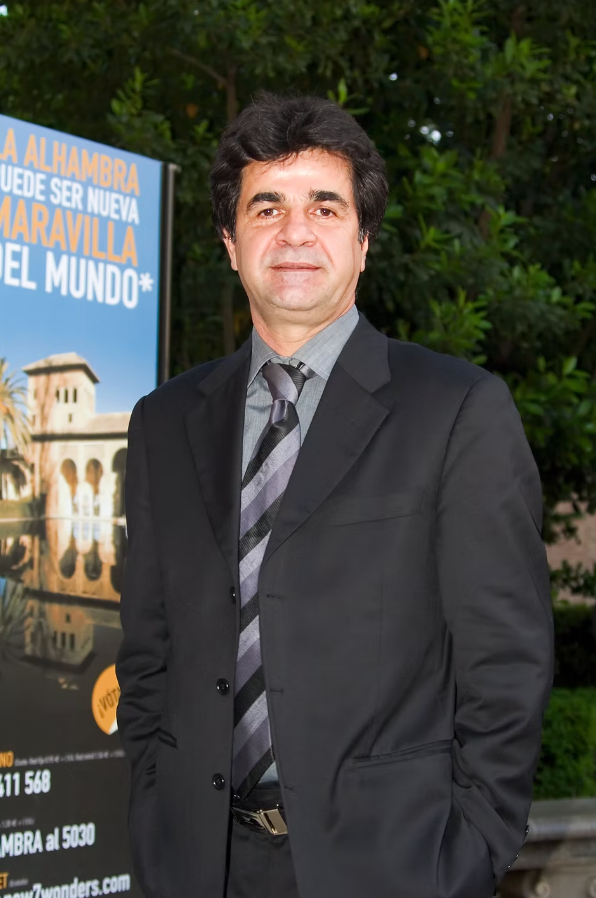 A man in a dark suit and striped tie stands outdoors, smiling with his hands in his pockets. Behind him is a sign with Spanish text and an image of a historic building, with green foliage in the background.