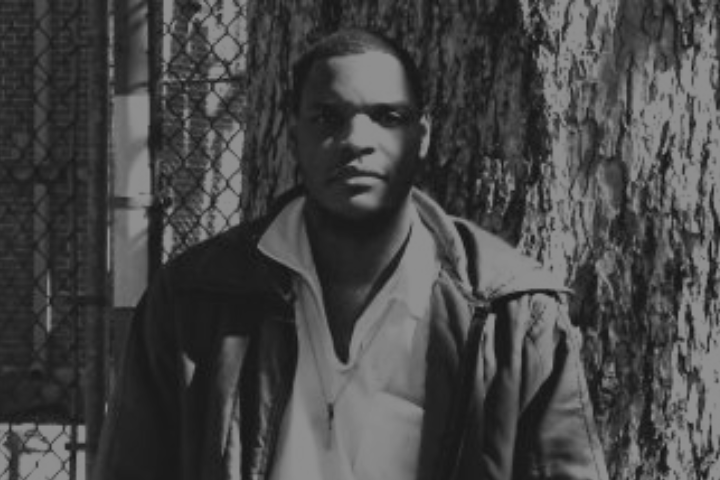 A man stands in front of a tree and a chain-link fence, wearing a jacket and glasses. The image is in black and white, with light and shadow creating contrast on his face and clothing.
