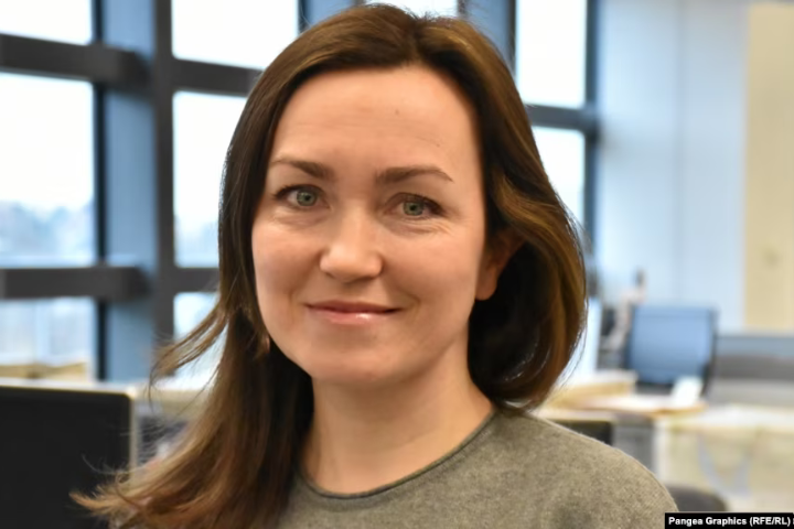 A woman with long brown hair and blue eyes, wearing a grey top, smiles slightly while standing in a bright office with large windows and blurred desks in the background.