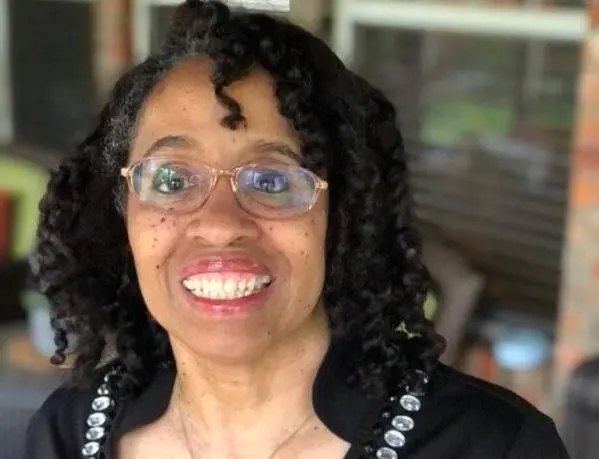 A smiling woman with glasses and curly hair wears a black top decorated with silver buttons. She is indoors, and the background is softly blurred.