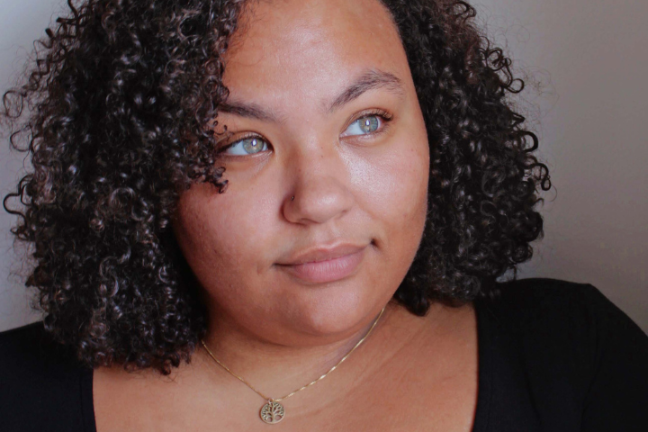 A person with curly dark hair and light-colored eyes looks slightly to the side, wearing a black top and a gold necklace with a round pendant, against a neutral background.