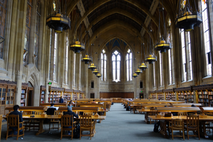 A grand library reading room with high arched ceilings, large windows, ornate chandeliers, long wooden tables, and bookshelves lining the walls. A few people are studying at the tables.