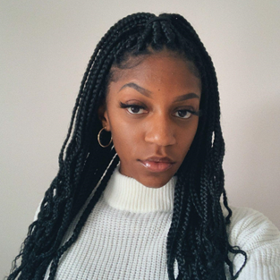 A woman with long black box braids, wearing a white ribbed turtleneck sweater and gold hoop earrings, poses in front of a plain light-colored background.
