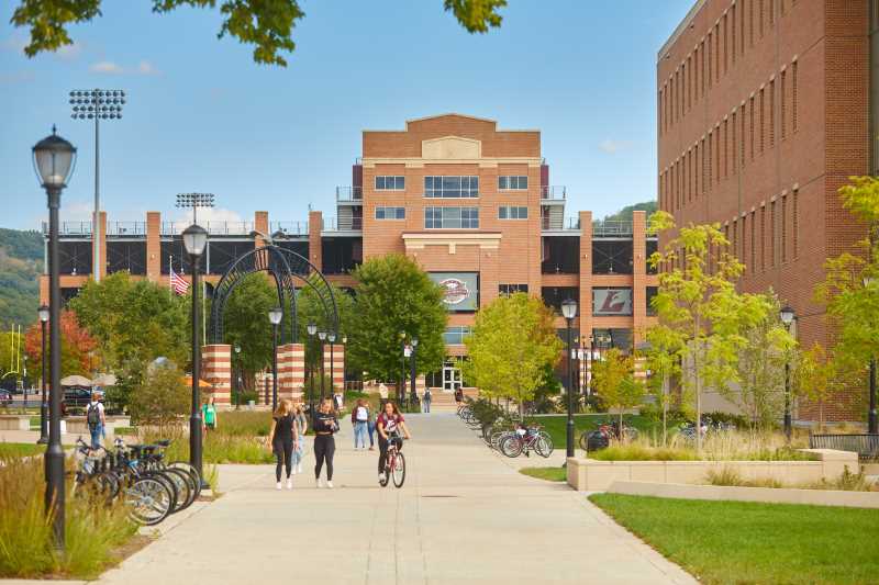 Students walk and ride bikes along a tree-lined campus pathway, with a brick academic building on the right and a sports stadium in the background under a blue sky.