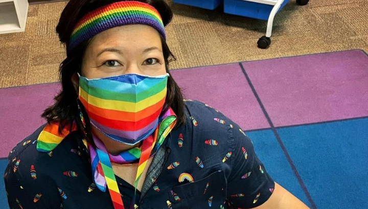 A person wearing a rainbow-striped face mask, headband, and lanyard sits on a colorful carpet. They have dark shoulder-length hair and are dressed in a navy shirt with rainbow patterns, showing pride and inclusivity.