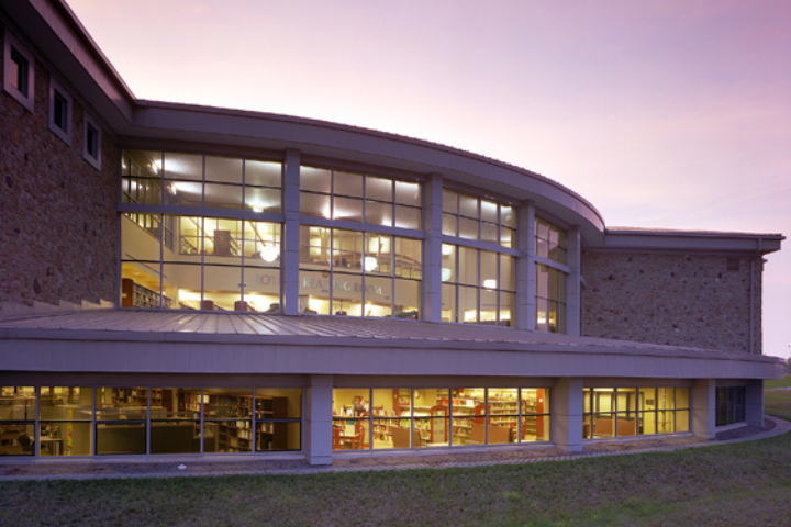 A modern building with large, curved glass windows is brightly lit from inside at dusk, showing multiple floors with visible bookshelves and people inside. The sky is purple and orange.
