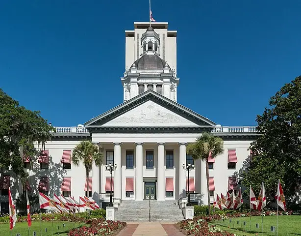 The front view of the Florida State Capitol building in Tallahassee, featuring white columns, a central dome, red-and-white window awnings, and multiple Florida state flags in the landscaped garden.