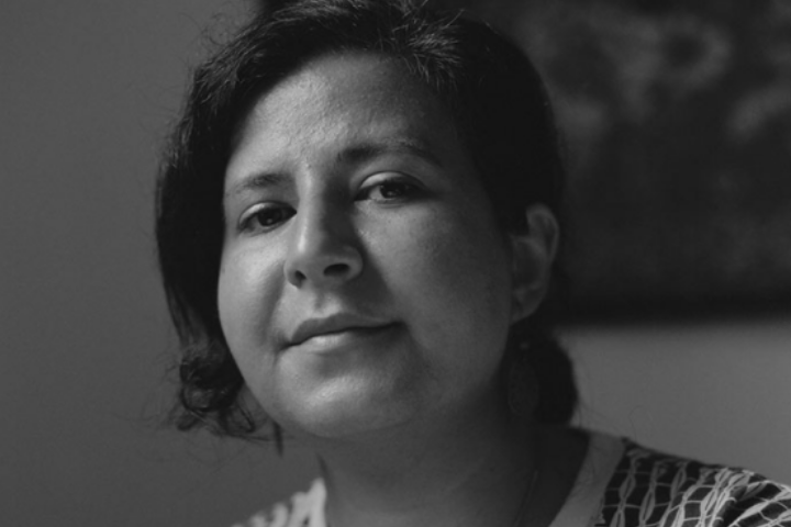 A person with short dark hair and a neutral expression looks slightly past the camera in a black-and-white portrait. The background is blurred, focusing attention on their face.