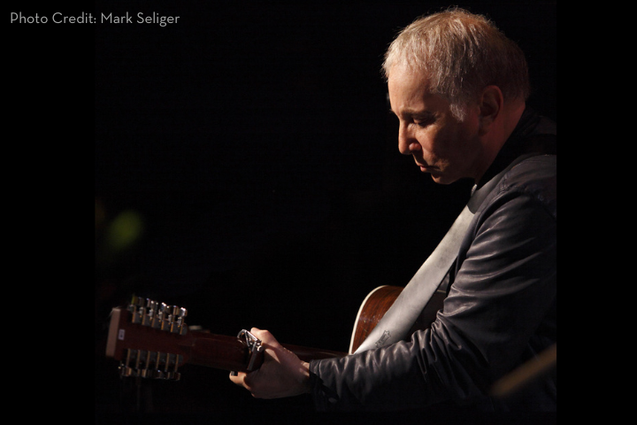 A man with short, light hair plays an acoustic guitar on stage, focused intently under dramatic lighting. He wears a dark jacket and the background is mostly black. Photo credit: Mark Seliger.