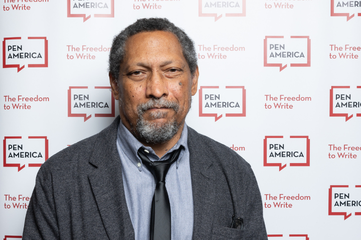 A man in a grey jacket and blue shirt stands in front of a PEN America step-and-repeat backdrop with the slogan The Freedom to Write in red text.