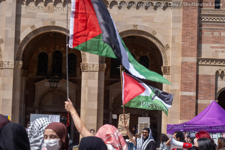 A group of people protest outside a brick building, holding Palestinian flags and signs. Some wear headscarves, and one sign reads STOP GENOCIDE. The scene is lively with various banners and tents.