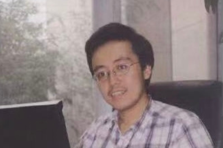 A young man with short dark hair and glasses, wearing a plaid shirt, sits indoors in front of a window with trees visible outside. He is looking slightly towards the camera and appears to be smiling.