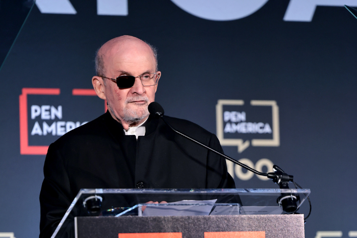 An older man wearing glasses and an eye patch stands at a podium, speaking into a microphone. He is dressed in a black jacket, with PEN America logos visible in the background.