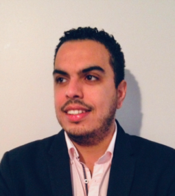 A man with short curly hair and facial hair, wearing a dark blazer over a light pink shirt, stands in front of a plain light-colored wall, looking slightly to his left and smiling.