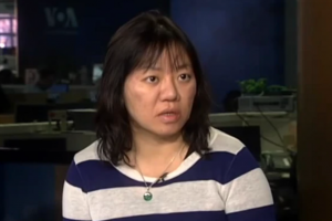 A woman with medium-length dark hair wearing a navy and white striped shirt and a green pendant necklace sits indoors, looking slightly to the side with a neutral expression.