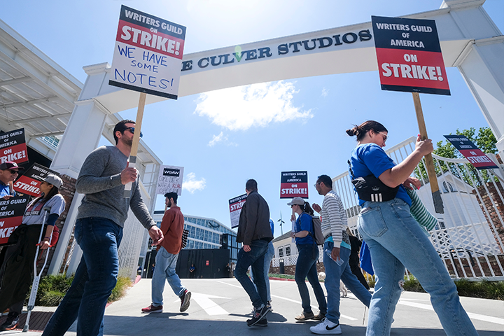 A group of people holding signs that read Writers Guild on Strike and We have some notes! picket outside E Culver Studios on a sunny day.