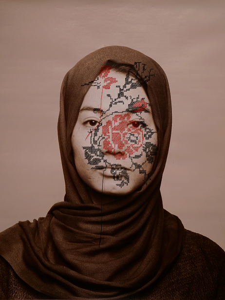 A woman in a brown hijab faces the camera. Her face is partially covered with an embroidered floral pattern, featuring red and black stitches, blending with her skin and expressionless gaze. The background is plain beige.