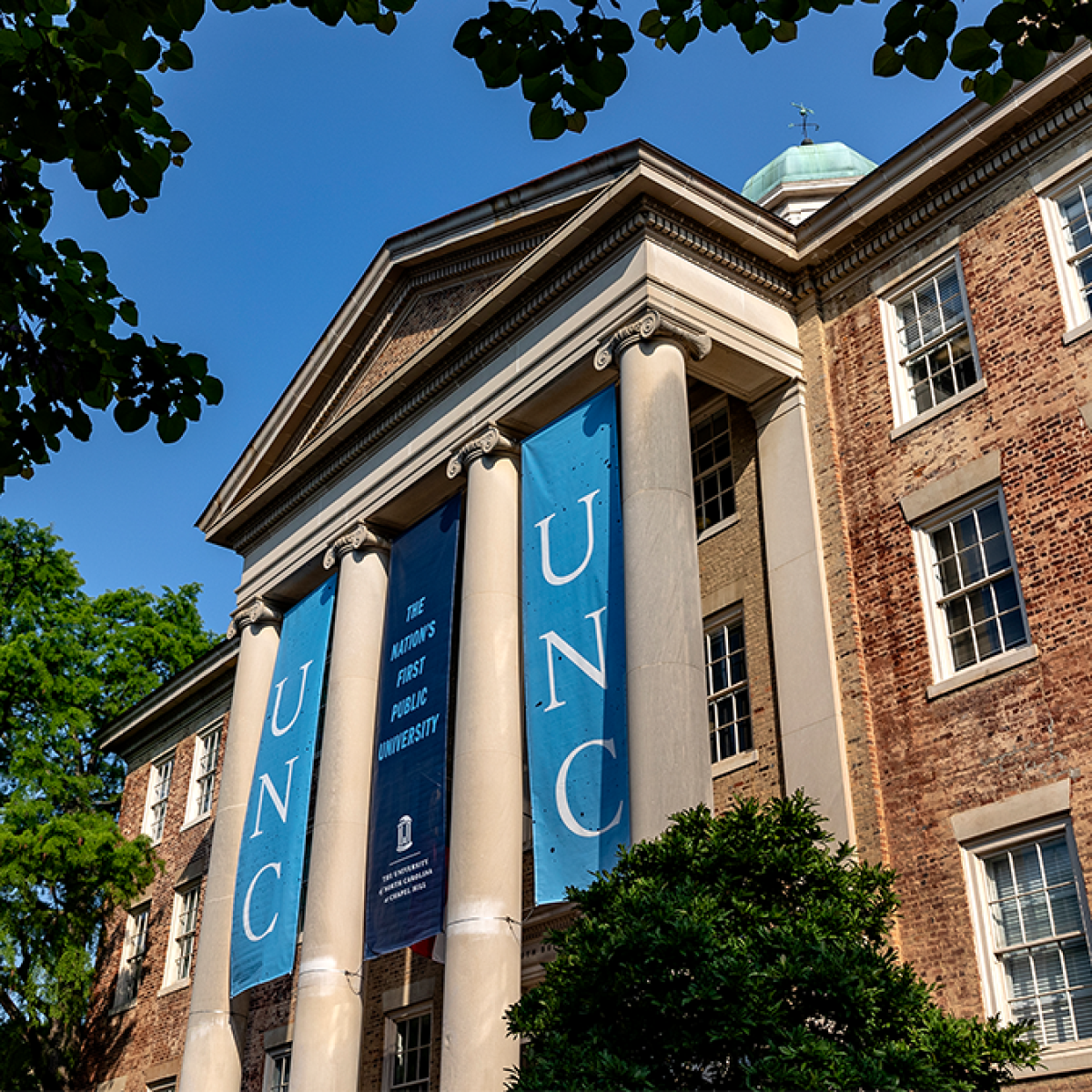 A historic brick university building with large white columns displays two vertical banners reading UNC and a central banner stating, The Nations First Public University, on a sunny day.