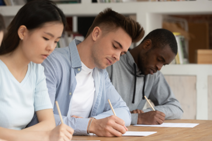 Three students sit at a table, focused on writing or taking a test, each holding a pencil. Bookshelves are visible in the background, suggesting a classroom or library setting.