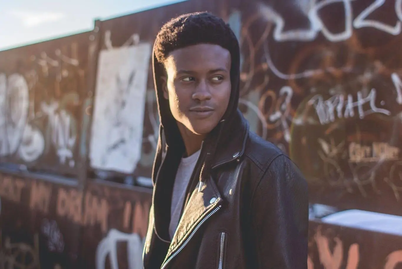 A young man wearing a black leather jacket and hoodie stands outdoors in front of a graffiti-covered wall, looking to the side with a thoughtful expression. The sunlight casts a warm glow on his face.
